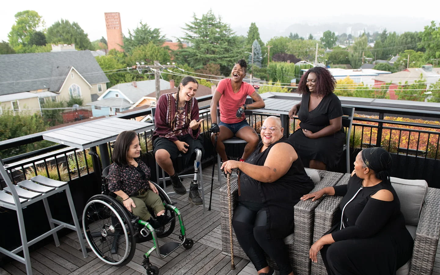 A group of disabled women — including someone using a wheelchair, someone with a prosthetic leg, and someone using a forearm crutch — smiling and talking together on an outdoor rooftop